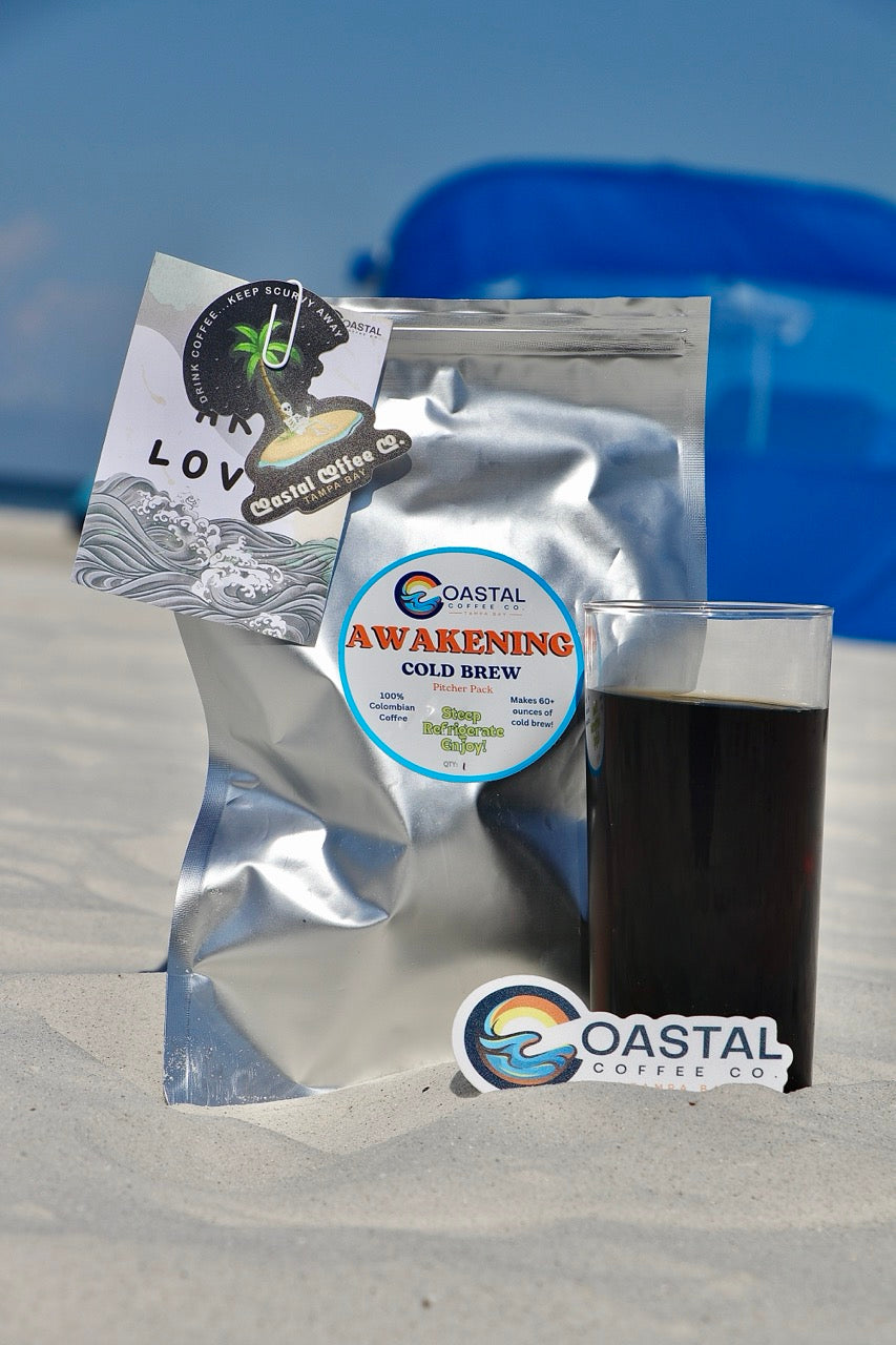 Awakening cold brew package at the beach sitting next to a glass of the cold brew and a Coastal Coffee Co. sticker.  